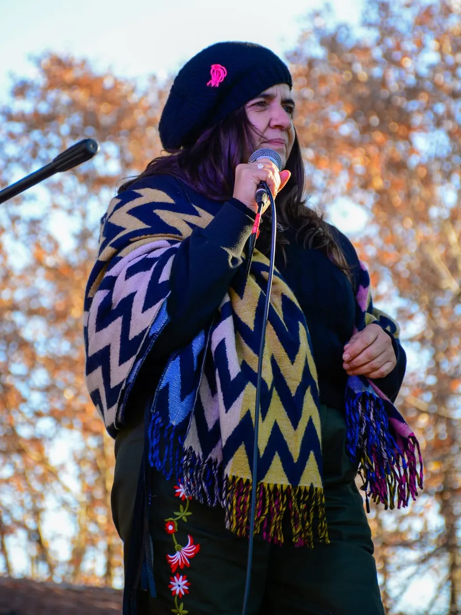 Lina cantando al micrófono con poncho de zigzag y boina negra entre árboles otoñales